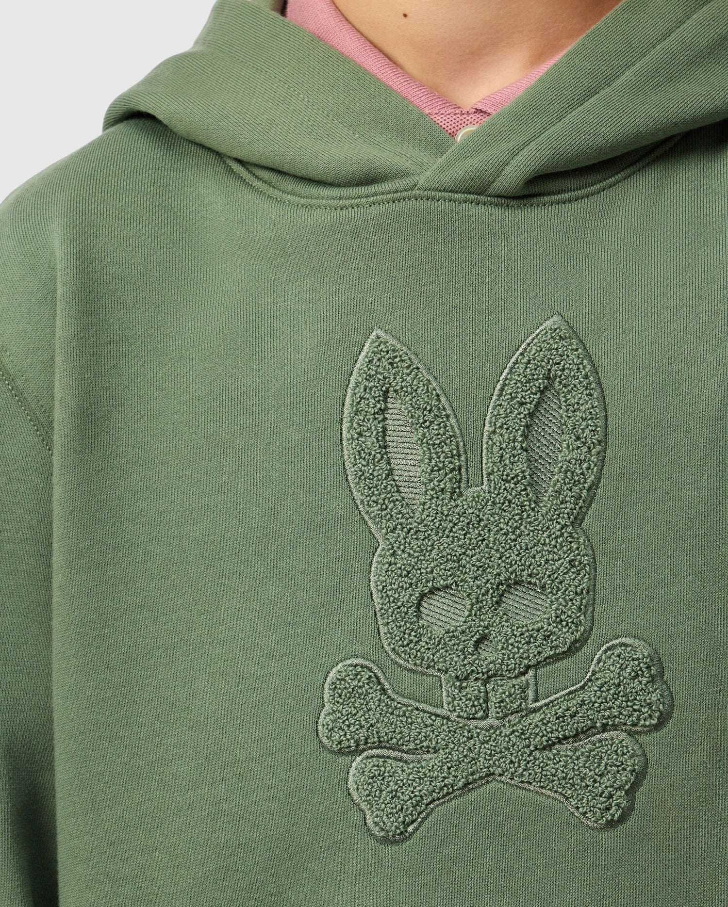 Close-up of a green KIDS PACIFIC CHENILLE HOODIE - B0H930D200 from Psycho Bunny, featuring chenille embroidery of a stylized bunny with long ears above two crossed bones. A hint of a pink shirt is visible underneath the hoodie.