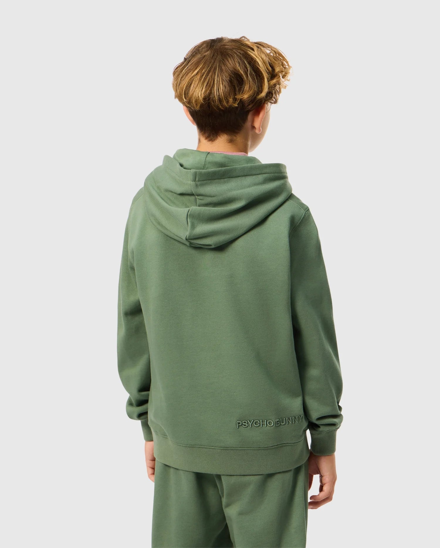 A young person with brown wavy hair is pictured facing away from the camera, wearing a green KIDS PACIFIC CHENILLE HOODIE - B0H930D200 from Psycho Bunny, paired with matching pants. The chenille embroidery of "Psycho Bunny" adorns the hem at the back. A simple gray background accentuates this official Psycho Bunny ensemble.