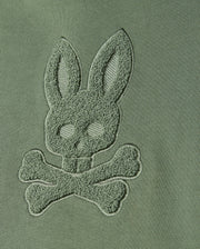 Featuring a close-up of green fabric adorned with chenille embroidery, the design presents a bunny head and crossed bones. This motif captures the essence of Psycho Bunny's distinct style, seamlessly blending into the fabric for a textured, raised monochromatic appearance—ideal for creating a masterpiece like the KIDS PACIFIC CHENILLE HOODIE (B0H930D200) from Psycho Bunny.