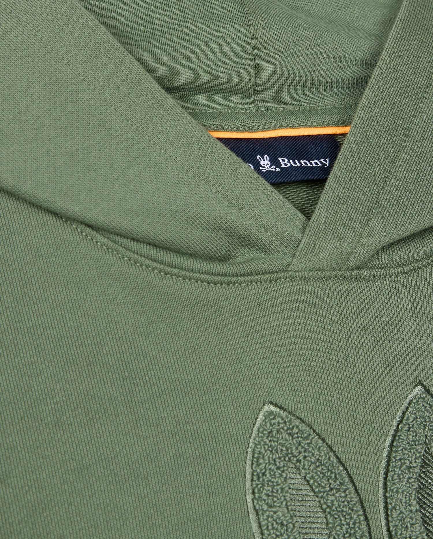 Close-up of the KIDS PACIFIC CHENILLE HOODIE in olive green French terry, featuring a subtle chenille leaf embroidery. Inside, a navy tag displays "Psycho Bunny." The fabric looks soft and textured.