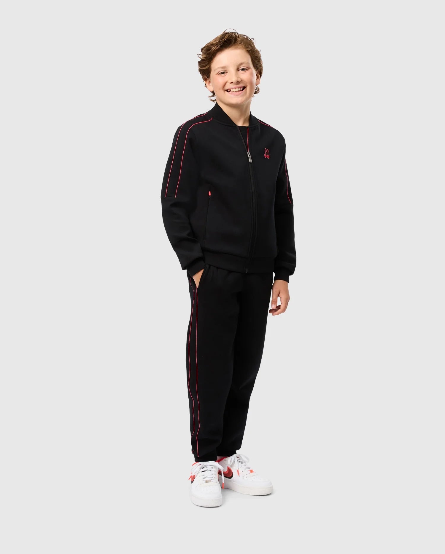 A young boy is standing and smiling, wearing a Psycho Bunny KIDS TRAVIS TRACK JACKET - B0J142D200 in black with red accents. The matching pants complete his regular fit tracksuit ensemble. Crafted from high-quality cotton-blend fabric, he pairs it perfectly with white sneakers featuring red details. The background is plain gray.