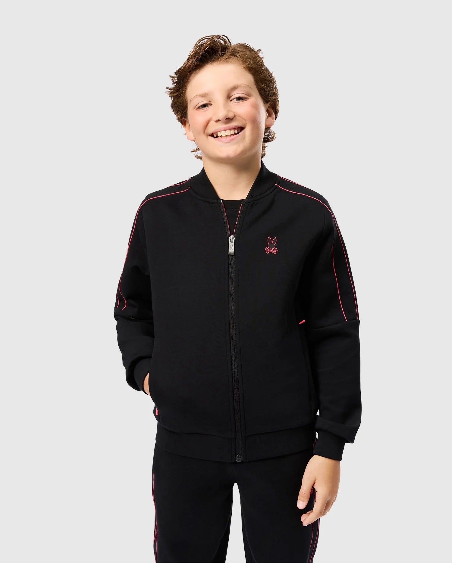 A smiling boy with short light brown hair is standing in front of a plain backdrop. He is wearing a black zip-up Psycho Bunny KIDS TRAVIS TRACK JACKET (B0J142D200) featuring red trim and a bunny logo on the chest, made from high-quality cotton-blend material. His hands are in his pockets, and he is dressed in matching regular fit black pants.