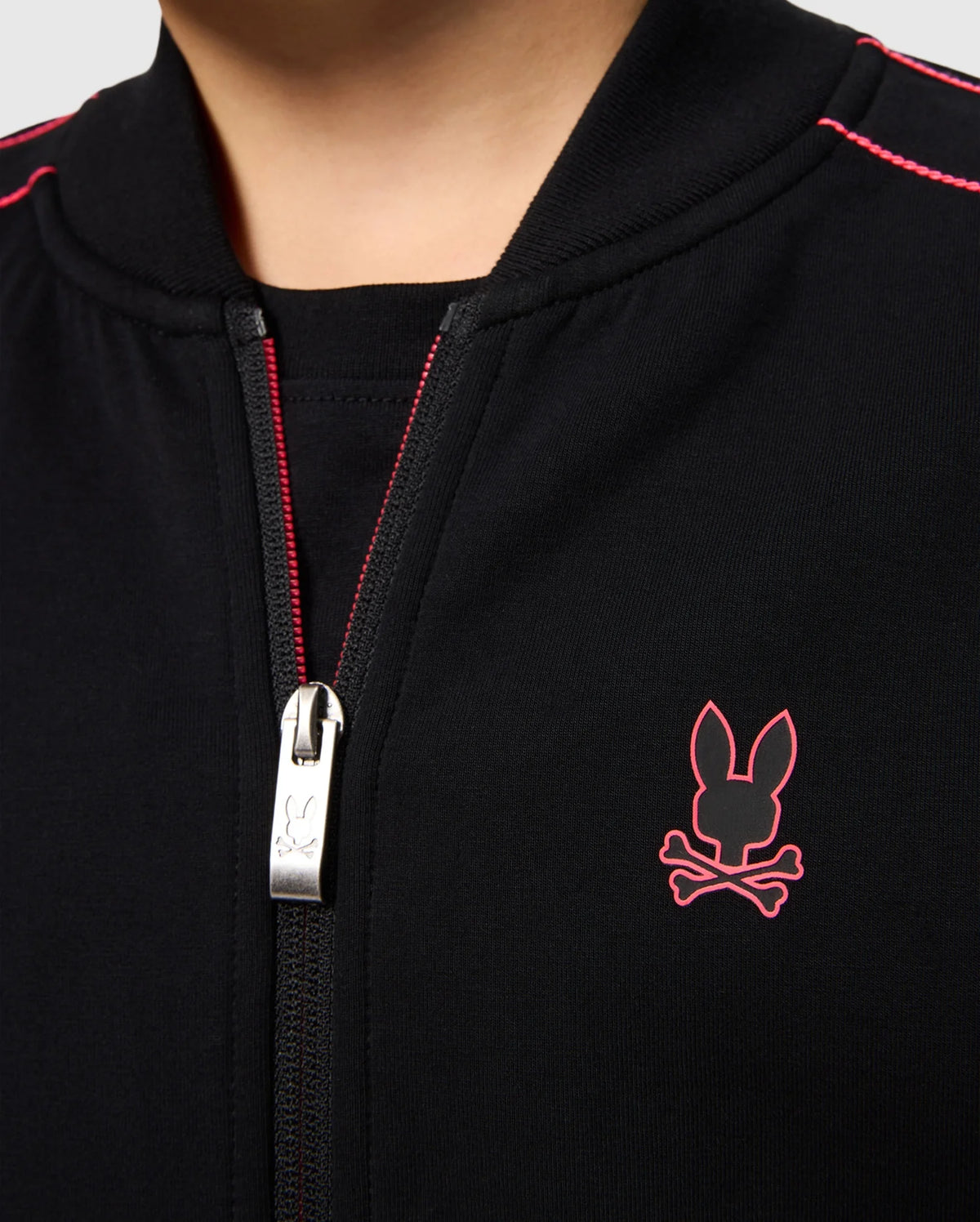 Close-up of a person wearing the KIDS TRAVIS TRACK JACKET - B0J142D200 by Psycho Bunny. The black regular fit jacket boasts a red stitched bunny logo and crossbones on the left side of the chest. Made from high-quality cotton-blend, it features a silver zipper with an engraved bunny emblem. Red stitching details accentuate the shoulders.