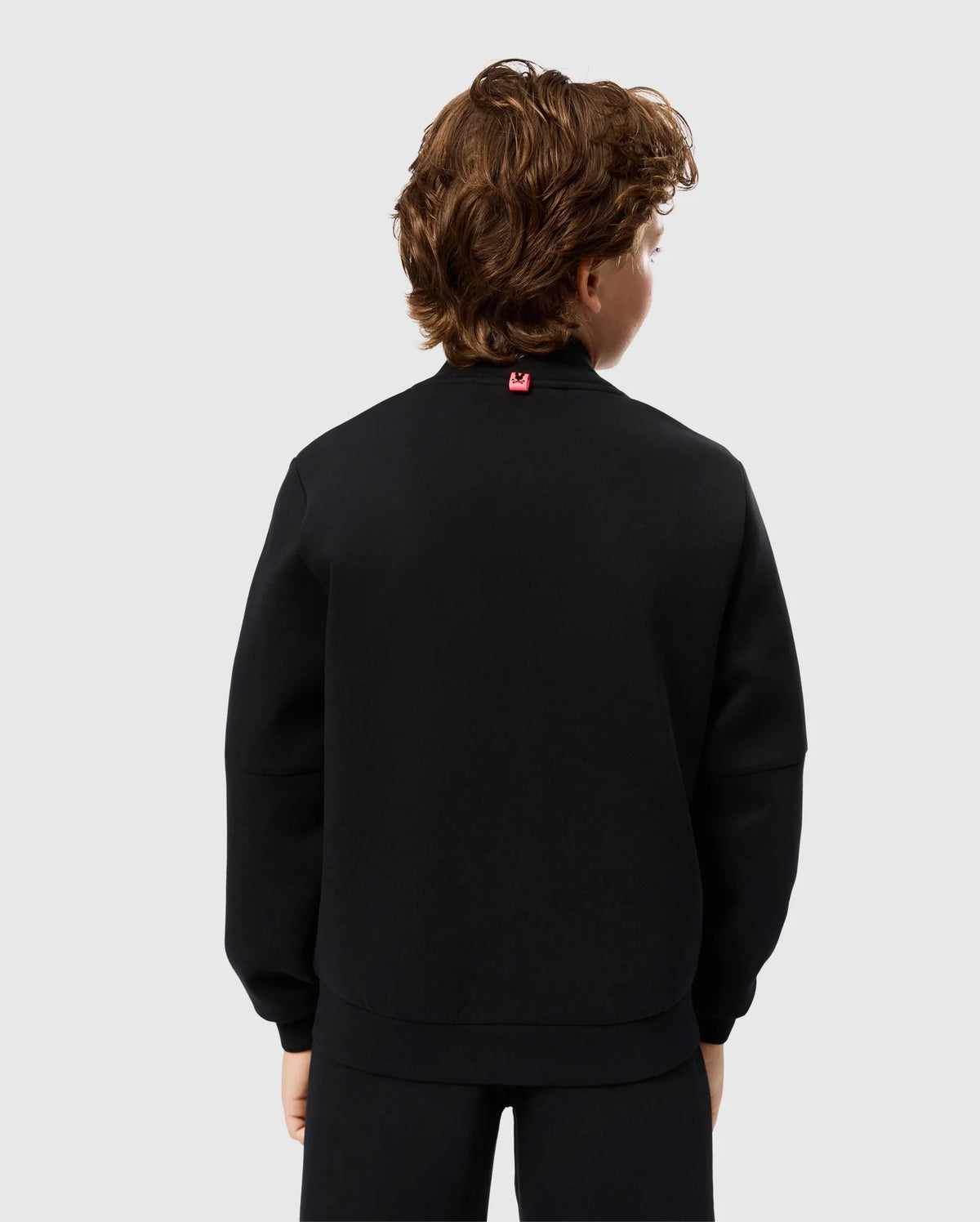 A person with curly brown hair is shown from the back, wearing a black long-sleeved Kids Travis Track Jacket by Psycho Bunny and black pants. The high-quality cotton-blend jacket, identified as model B0J142D200, features a small red tag at the back of the neck. The background is plain and light gray.