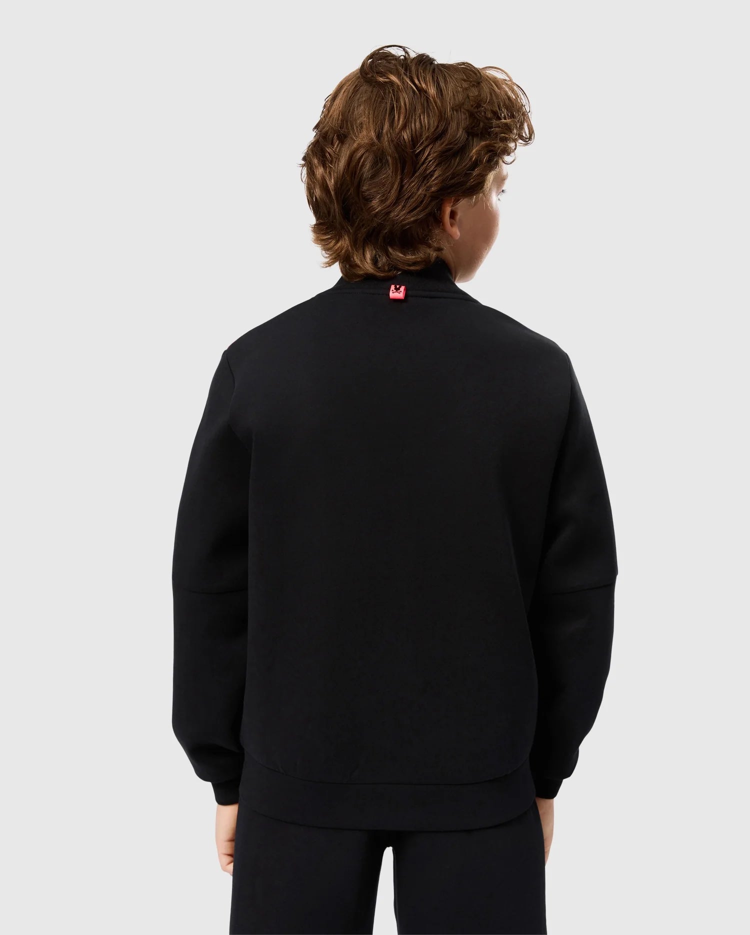 A person with curly brown hair is shown from the back, wearing a black long-sleeved Kids Travis Track Jacket by Psycho Bunny and black pants. The high-quality cotton-blend jacket, identified as model B0J142D200, features a small red tag at the back of the neck. The background is plain and light gray.