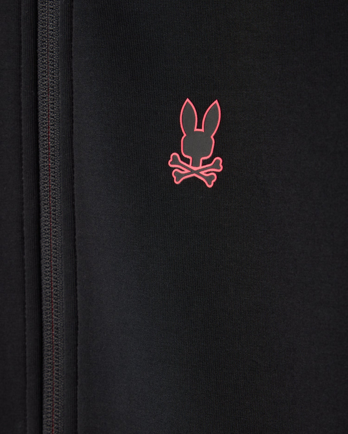A close-up of the KIDS TRAVIS TRACK JACKET - B0J142D200 by Psycho Bunny, designed in a regular fit and crafted from high-quality cotton-blend fabric with a zipper on the left side. It features a small pink logo of a rabbit's head with crossbones underneath on the right side.
