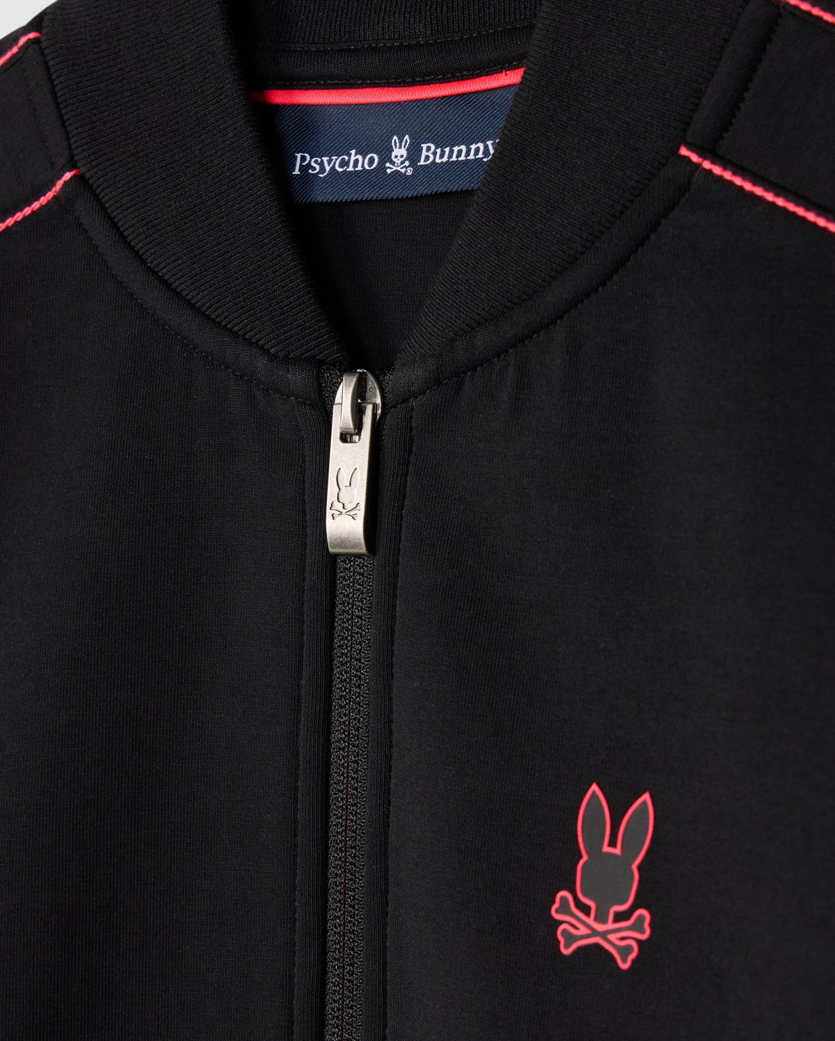 Close-up of a black Kids Travis Track Jacket (B0J142D200) by Psycho Bunny, featuring red stitching and a small red emblem of a bunny with crossbones. Made from high-quality cotton-blend, it includes a zipper pull adorned with the same bunny design. Inside the collar, a label reads "Psycho Bunny," ensuring its regular fit.