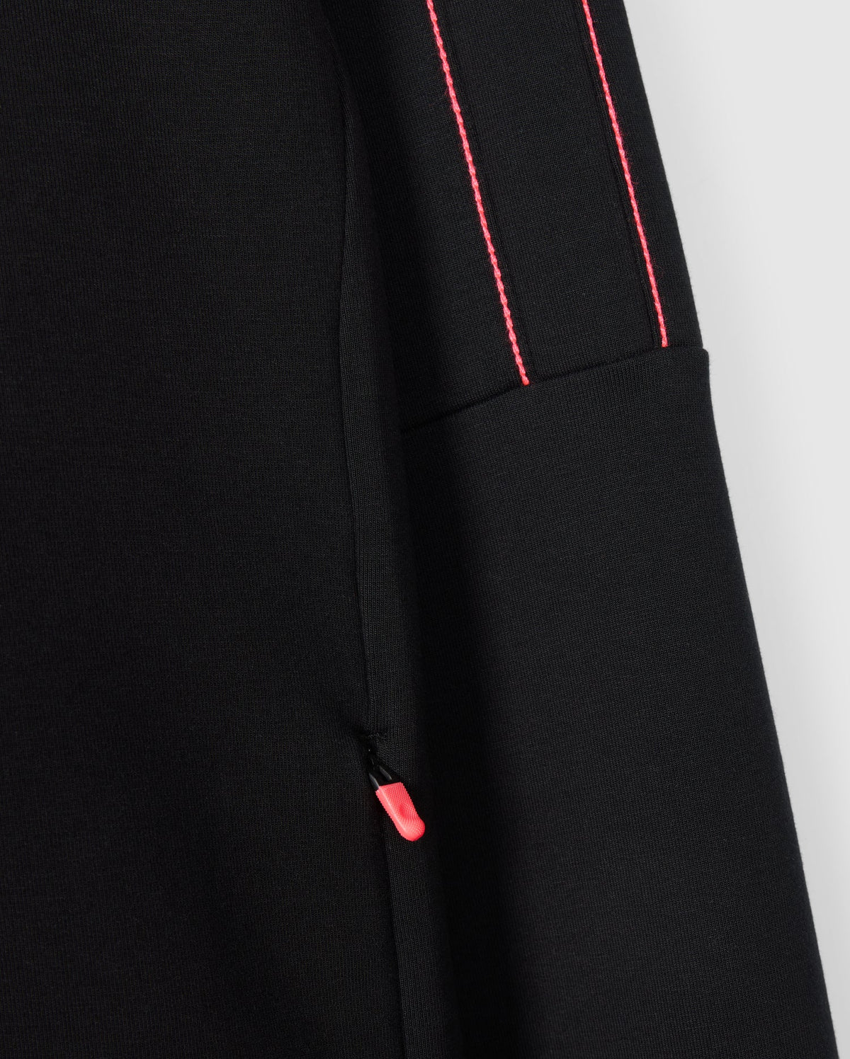 Close-up of the Psycho Bunny Kids Travis Track Jacket (B0J142D200), crafted from a high-quality cotton-blend fabric. The jacket features a small pink zipper pull on the pocket seam, and the sleeve showcases two thin, parallel pink stitches running vertically, adding a contrasting detail to the predominantly black fabric. The texture appears smooth and slightly stretchy.