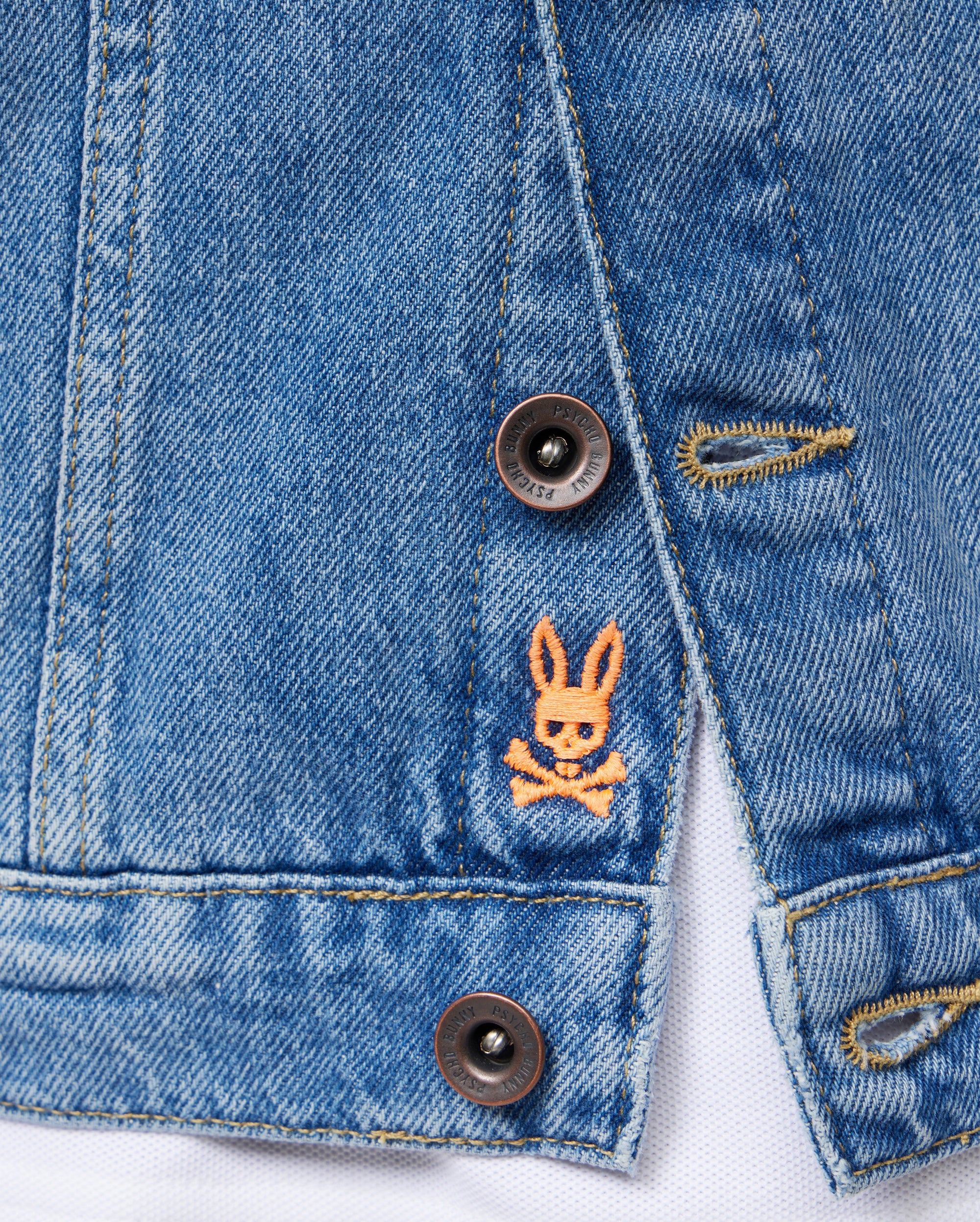 Close-up of a section of the Psycho Bunny KIDS PATHE DENIM JACKET - B0J741X1CO made from upcycled cotton, featuring two buttons and a small embroidered logo of an orange bunny head with crossed bones beneath it. The stitching and buttonholes are also visible, providing texture to the fabric.
