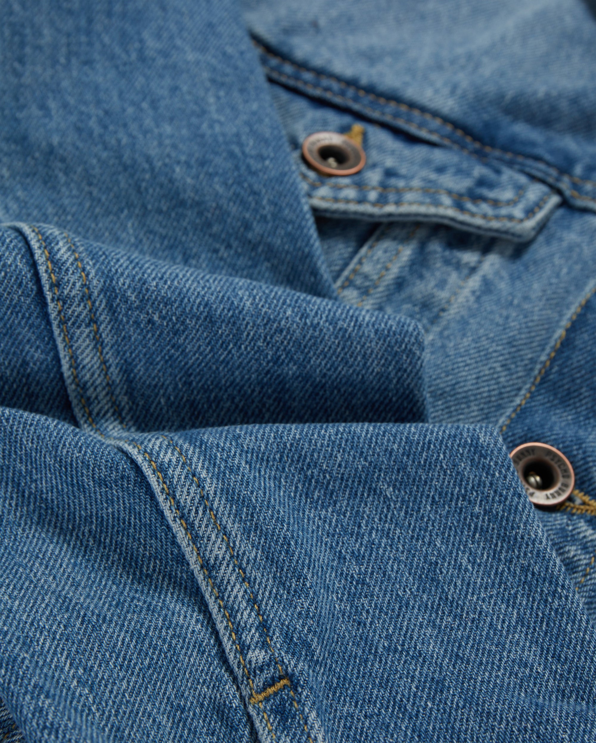 Close-up of blue denim fabric from a Psycho Bunny KIDS PATHE DENIM JACKET - B0J741X1CO, showing detailed stitching and two metal buttons. The fabric is folded, creating soft creases and highlighting the texture of the material. The image captures the classic look of upcycled cotton denim, emphasizing its weave and durability.