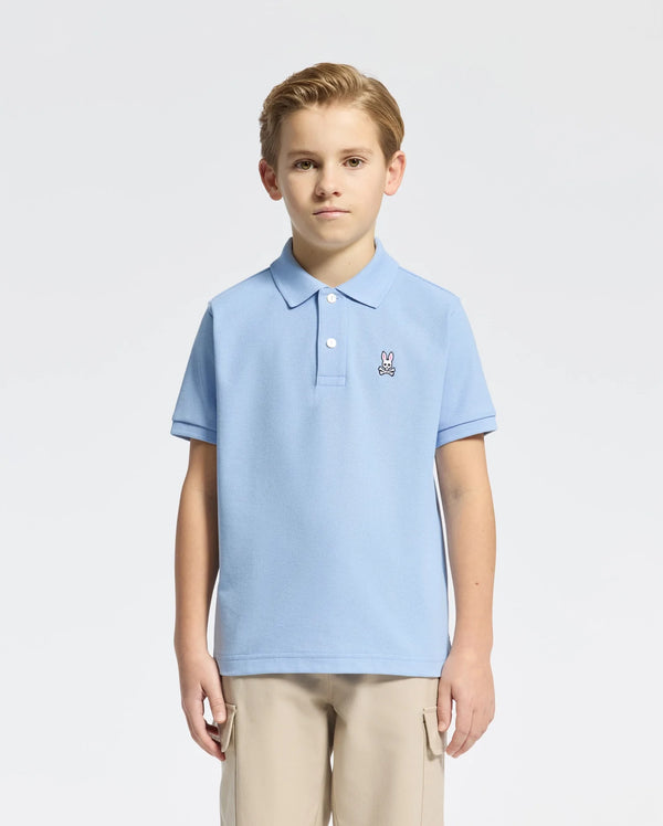 A young boy with short light brown hair stands against a white background, wearing Psycho Bunny's Kids Classic Pique Polo (B0K001B200), a light blue Pima cotton shirt with an embroidered bunny logo on the chest, and beige pants, his hands resting at his sides.
