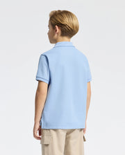A young child with short blond hair faces away, wearing a Psycho Bunny Kids Classic Pique Polo in light blue and beige pants. The plain, light-colored background highlights the outfit's simplicity.