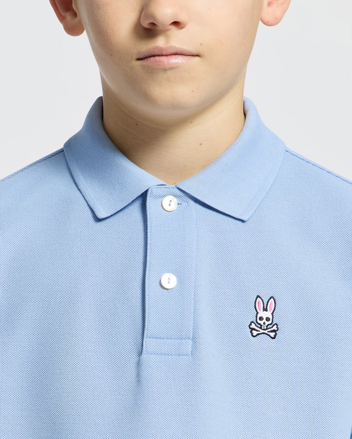 A person wearing the Psycho Bunny KIDS CLASSIC PIQUE POLO - B0K001B200 with an embroidered pink bunny and crossed bones, showcased in a cropped light blue image focusing on the lower face, neck, and upper torso.