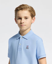 A boy with short brown hair wears a light blue KIDS CLASSIC PIQUE POLO by Psycho Bunny, adorned with a small embroidered bunny logo on the chest, set against a plain white background.