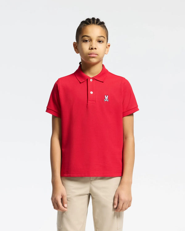 A young person with braided hair wears a red Psycho Bunny KIDS CLASSIC PIQUE POLO SHIRT (B0K001CRPC) featuring a small rabbit emblem on the chest, paired with beige pants, set against a white background—ideal for back-to-school style.
