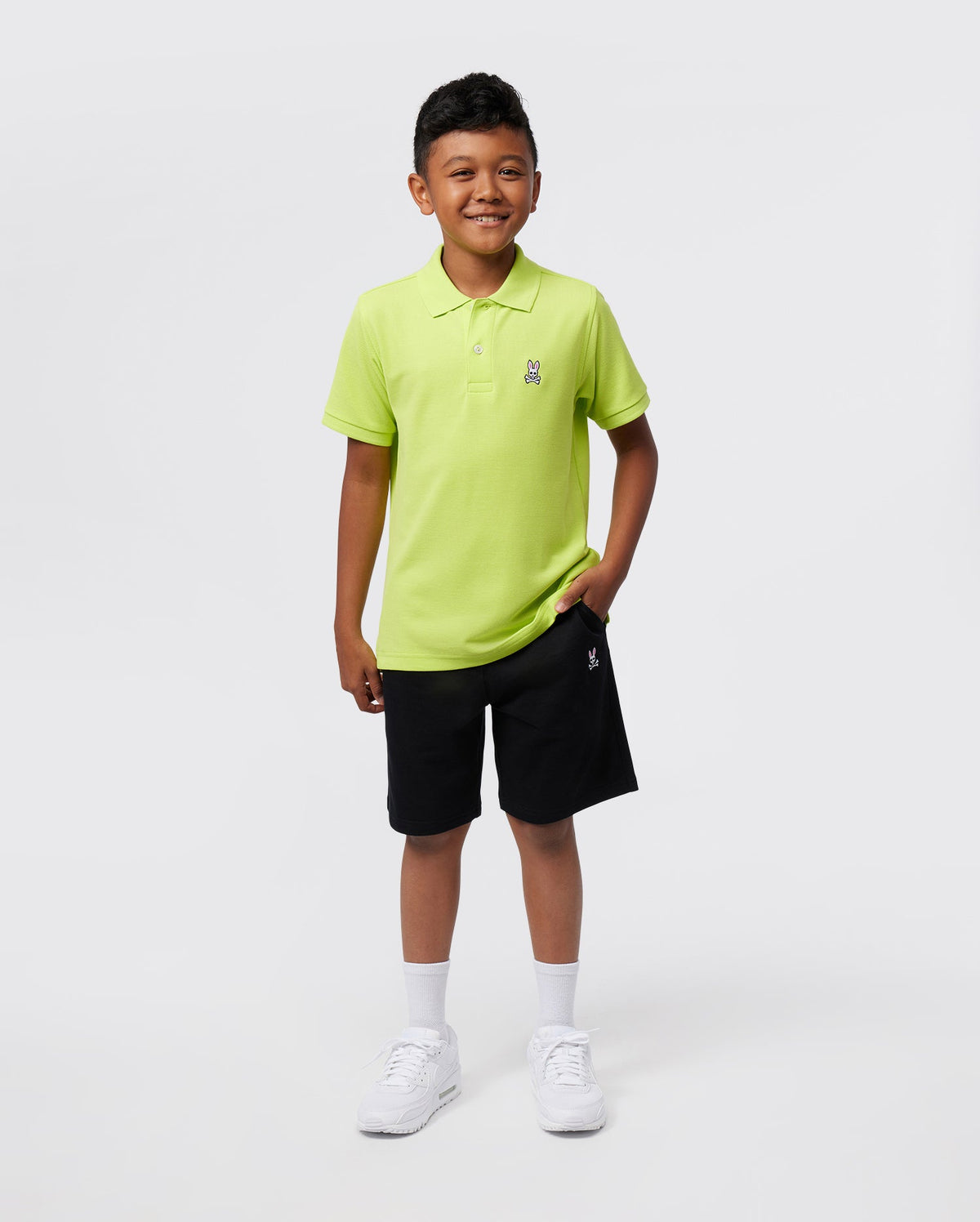 A young boy smiles against a plain background, wearing the Psycho Bunny KIDS CLASSIC POLO - B0K001CRPC in bright green, crafted from soft Pima cotton. He's paired it with black shorts, white socks, and white sneakers, casually placing one hand in his pocket.