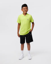 A young boy smiles against a plain background, wearing the Psycho Bunny KIDS CLASSIC POLO - B0K001CRPC in bright green, crafted from soft Pima cotton. He's paired it with black shorts, white socks, and white sneakers, casually placing one hand in his pocket.