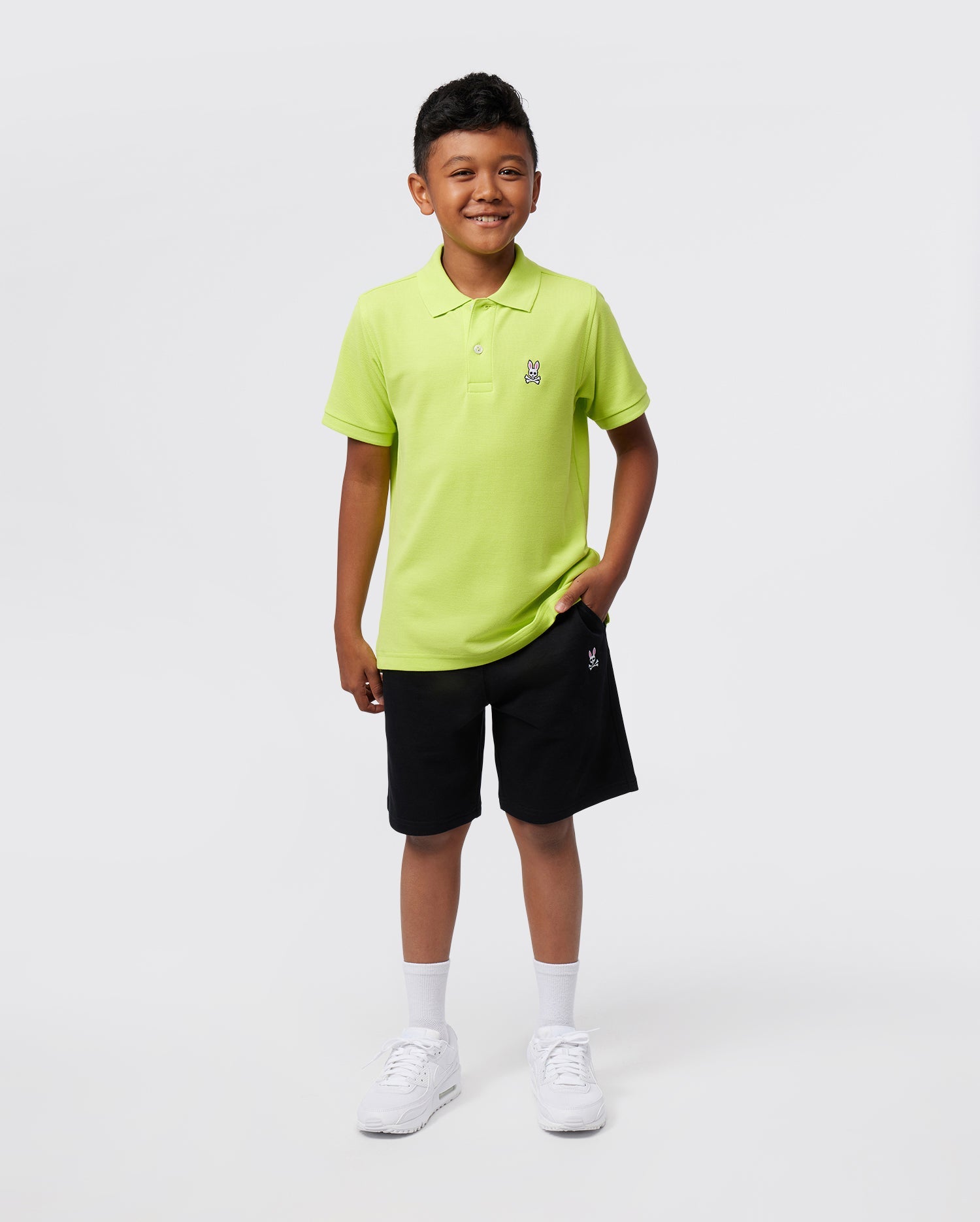 A young boy smiles against a plain background, wearing the Psycho Bunny KIDS CLASSIC POLO - B0K001CRPC in bright green, crafted from soft Pima cotton. He's paired it with black shorts, white socks, and white sneakers, casually placing one hand in his pocket.