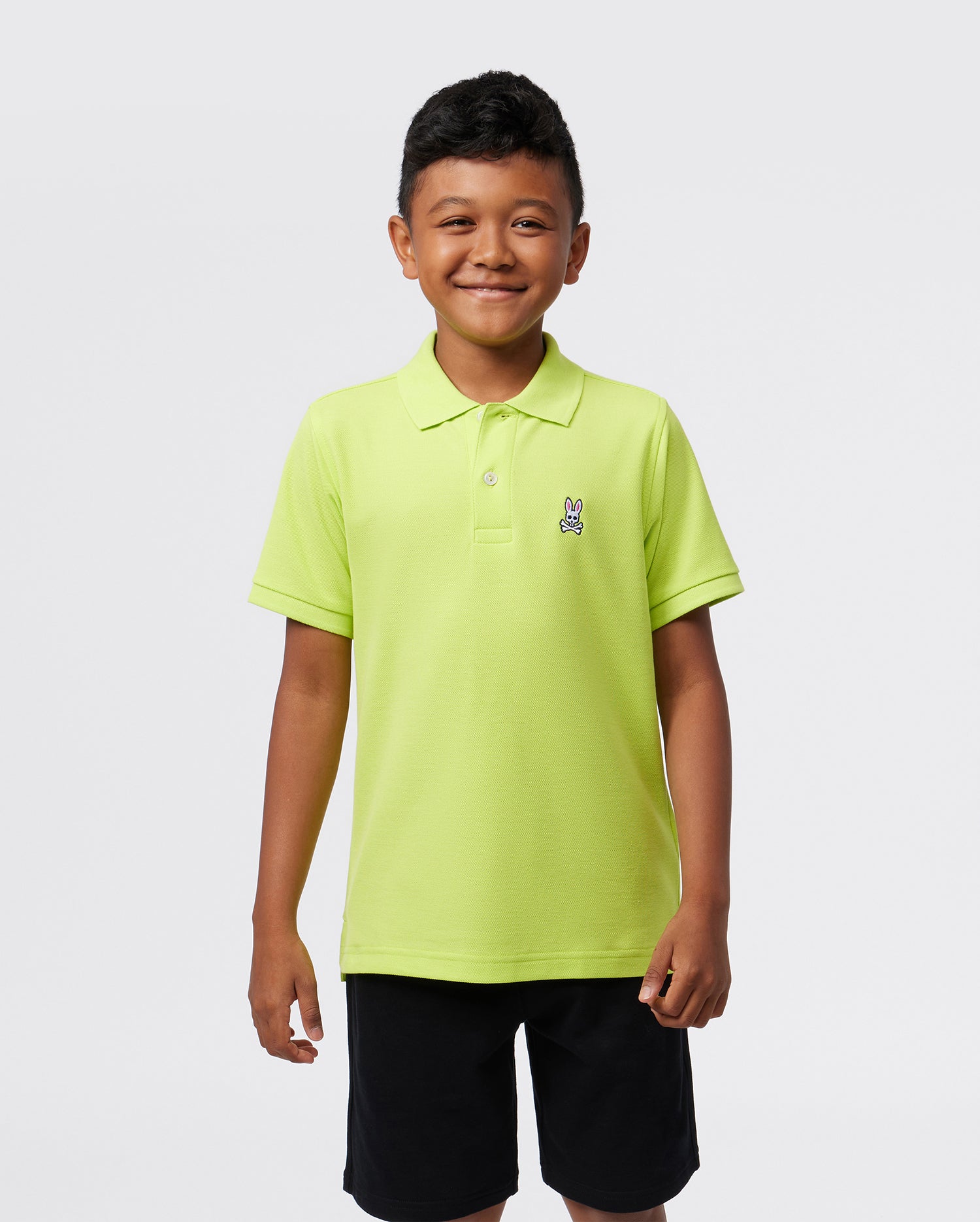 A smiling child, confident against a plain background, wears the Psycho Bunny KIDS CLASSIC POLO - B0K001CRPC in bright green. The regular fit Pima cotton piqué features a small emblem on the left side, enhancing their cheerful appearance.