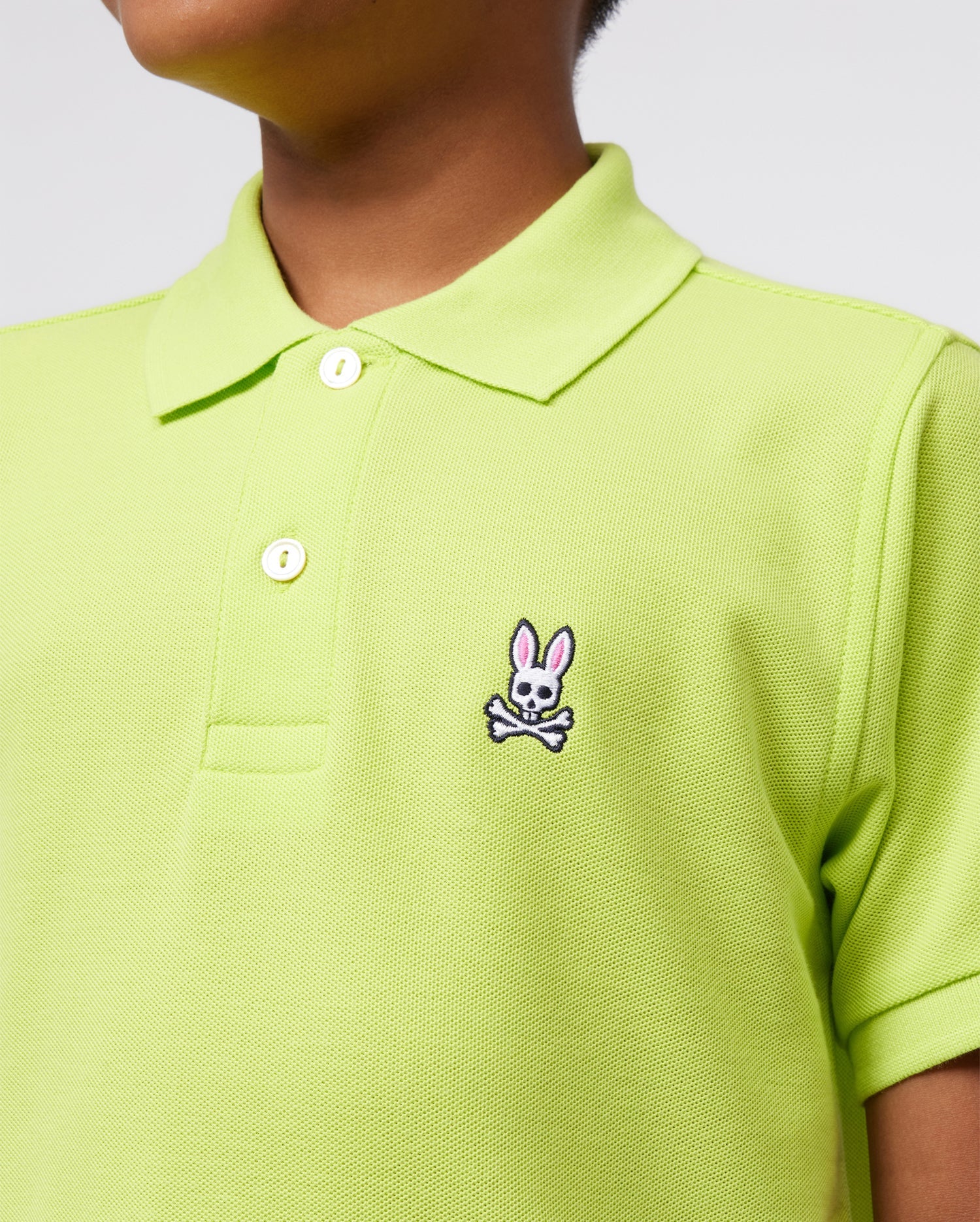 Someone is wearing a lime green Psycho Bunny Kids Classic Polo made from soft Pima cotton. It features a small pink and white bunny logo above crossed bones on the chest, has buttons, a collar, and a regular fit in front of a plain background.
