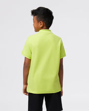 A young boy stands with his back to the camera, wearing a Psycho Bunny KIDS CLASSIC POLO - B0K001CRPC in light green made from soft Pima cotton, paired with black shorts against a plain white background.