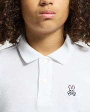 A curly-haired person wears a KIDS CLASSIC PIQUE POLO SHIRT by Psycho Bunny, ideal for back-to-school outfits. This white shirt stands out with its unique chest logo—a pink-eared rabbit above crossed bones.