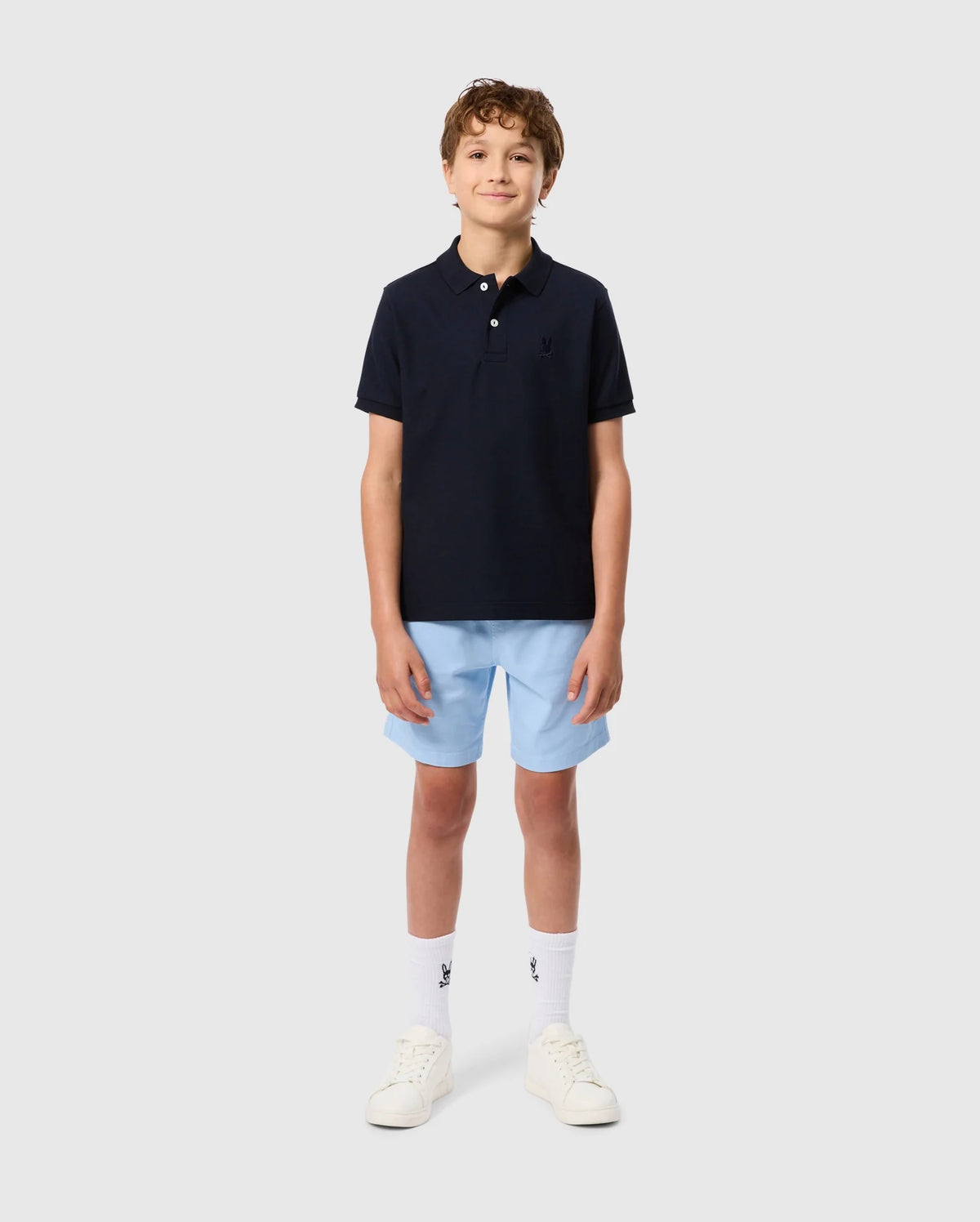 A young person stands against a plain background, wearing the Psycho Bunny KIDS CLASSIC PIQUE POLO SHIRT - B0K021AR00 with an embroidered chest Bunny, paired with light blue shorts, white socks, and white sneakers. Their hands rest by their sides as they smile casually.
