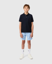 A young person stands against a plain background, wearing the Psycho Bunny KIDS CLASSIC PIQUE POLO SHIRT - B0K021AR00 with an embroidered chest Bunny, paired with light blue shorts, white socks, and white sneakers. Their hands rest by their sides as they smile casually.