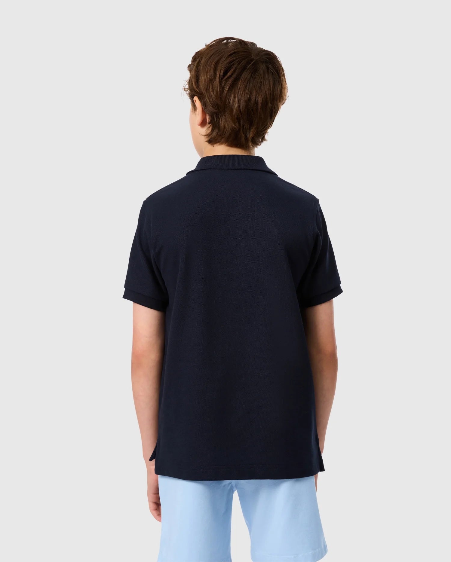 A young person with brown hair is wearing a Psycho Bunny KIDS CLASSIC PIQUE POLO SHIRT - B0K021AR00 in classic dark navy, paired with light blue shorts, standing with their back to the camera. The embroidered chest bunny adds a playful touch against the plain light gray background.