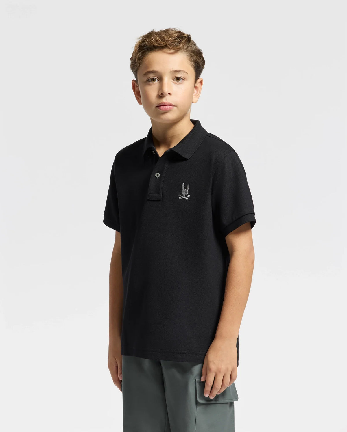 A young boy wearing a Psycho Bunny KIDS ALEXANDER PIQUE POLO - B0K101E200, which features a small embroidered logo on the chest, stands against a plain white background. With short brown hair, he looks directly at the camera while pairing the polo with gray pants, creating a fashion-forward vibe in kids' wear.