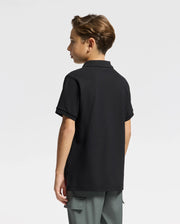 A young person with short brown hair is standing and facing away from the camera, wearing a Psycho Bunny Kids Alexander Pique Polo in black and gray pants against a plain white background.
