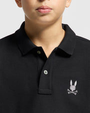 A person is wearing a Kids Alexander Pique Polo in black from Psycho Bunny, with two buttons undone, featuring a small embroidered bunny skull and crossbones logo on the left side. The plain background accentuates its fashion-forward design, making it ideal for any trendy occasion.