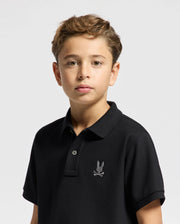 A young boy with short brown hair is wearing the KIDS ALEXANDER PIQUE POLO - B0K101E200 from Psycho Bunny, a black Pima cotton polo featuring a gray embroidered logo of a rabbit skull and crossbones. The plain white background highlights the high-quality kids' polo in all its charming simplicity.