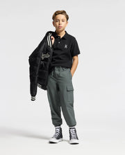 A young boy is wearing a Psycho Bunny Kids Alexander Pique Polo in black, matched with gray cargo pants and stylish black high-top sneakers. He casually drapes a bomber jacket over his shoulder against a plain white background.