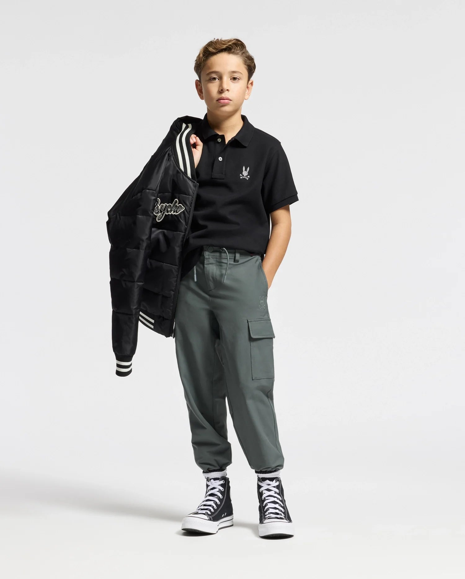 A young boy is wearing a Psycho Bunny Kids Alexander Pique Polo in black, matched with gray cargo pants and stylish black high-top sneakers. He casually drapes a bomber jacket over his shoulder against a plain white background.