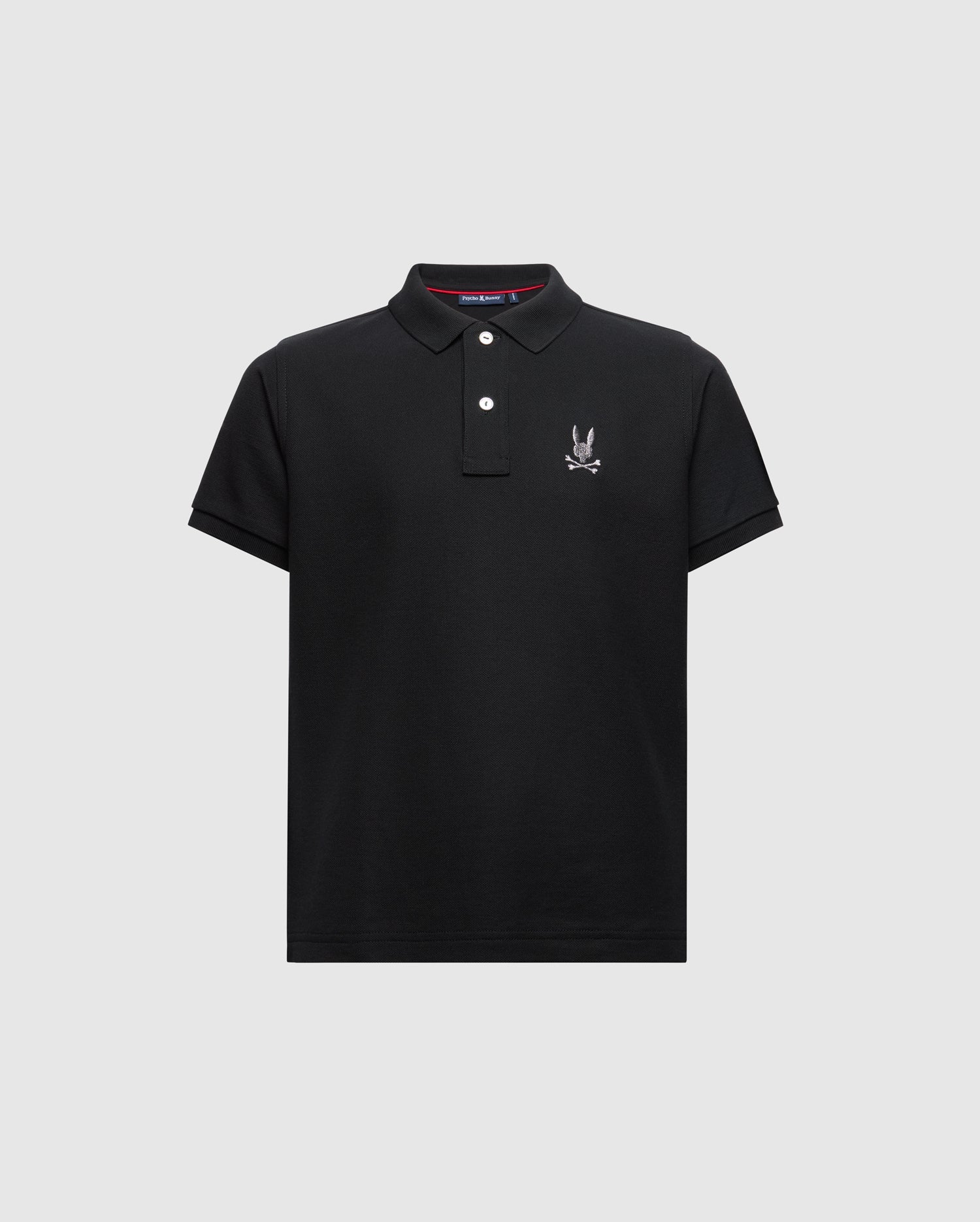 Introducing the KIDS ALEXANDER PIQUE POLO - B0K101E200 from Psycho Bunny. This fashionable black polo for kids features short sleeves and a buttoned collar, adorned with an embroidered logo showcasing a cartoon rabbit with crossed bones on the chest, elegantly set against a light gray background.