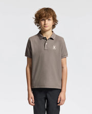 A young person with curly hair is wearing a stylish Psycho Bunny Kids Alexander Pique Polo, crafted from premium Pima cotton. The brown shirt, adorned with a small white logo on the chest, pairs perfectly with black pants as they stand against a plain white background.