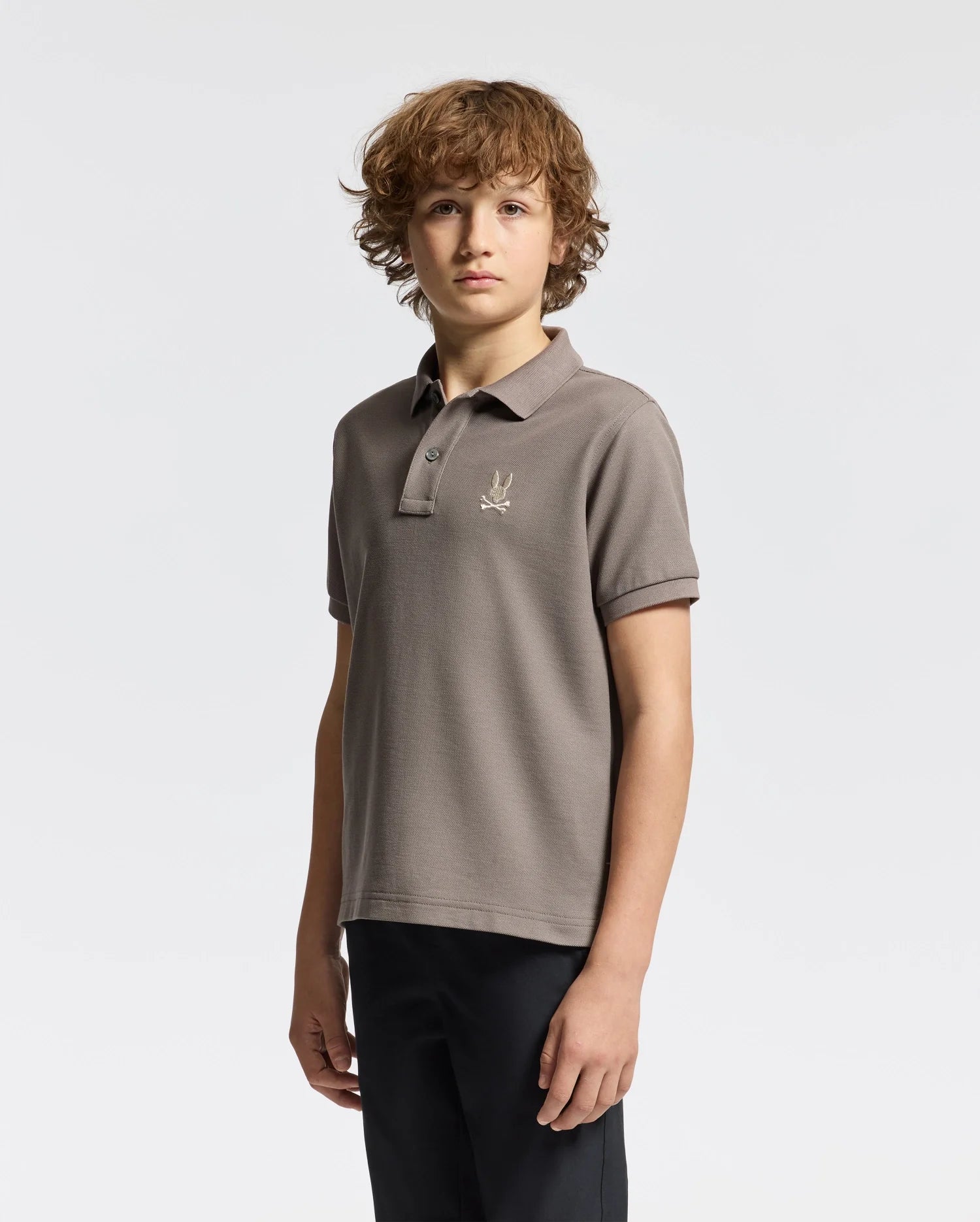 A young person with curly hair is wearing a gray KIDS ALEXANDER PIQUE POLO - B0K101E200 by Psycho Bunny, featuring an embroidered logo on the chest, paired with black pants, creating a stylish and modern look against a plain white background.