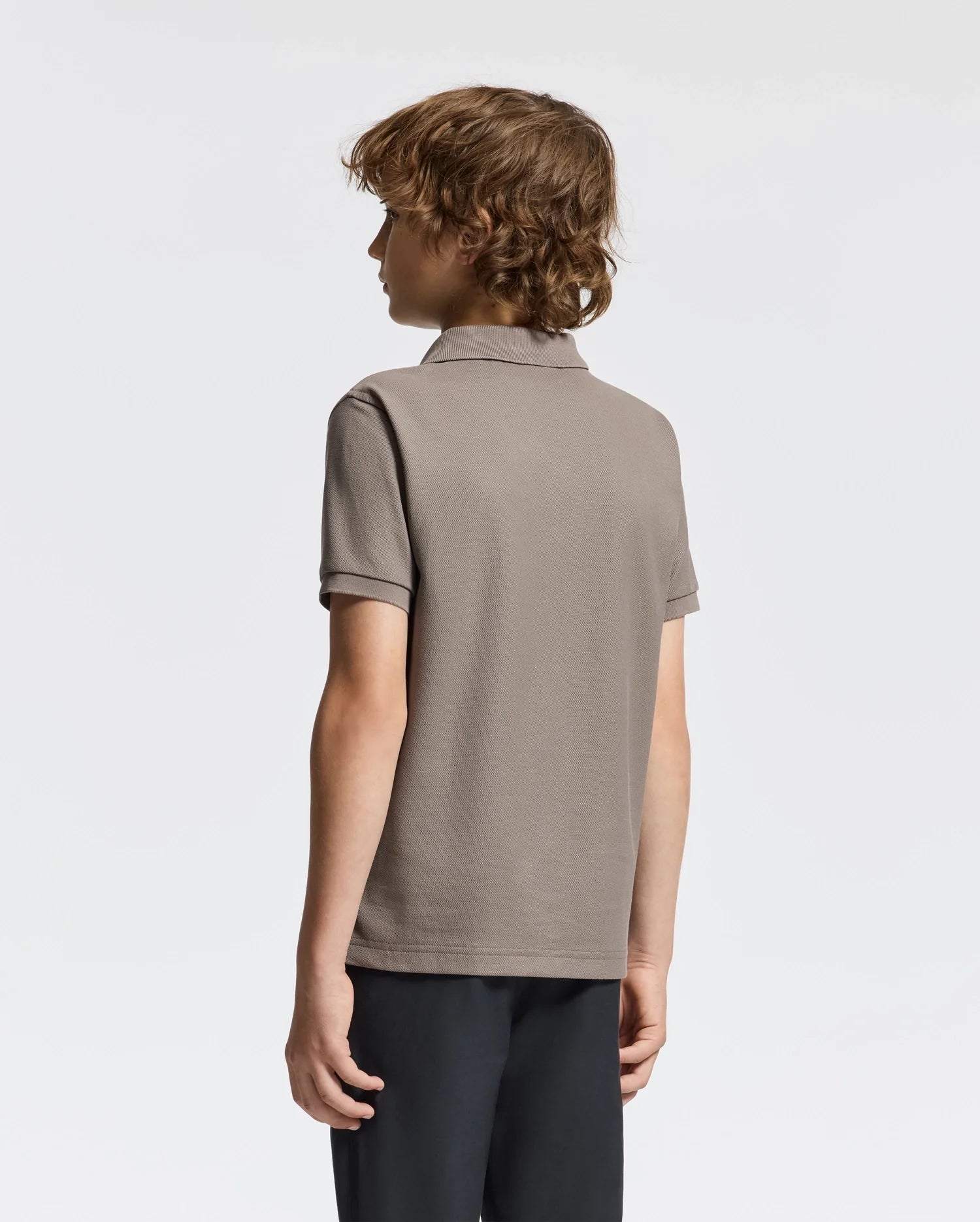 A person with curly hair stands facing away from the camera, highlighting their fashion-forward KIDS ALEXANDER PIQUE POLO by Psycho Bunny. The taupe short-sleeve shirt, made from soft Pima cotton, pairs effortlessly with dark pants against a light neutral background.