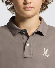 A person wearing a taupe KIDS ALEXANDER PIQUE POLO - B0K101E200 by Psycho Bunny, which features an embroidered rabbit skull and crossbones logo on the chest. This shirt is made from soft Pima cotton with two buttons and is depicted in close-up against a plain background.