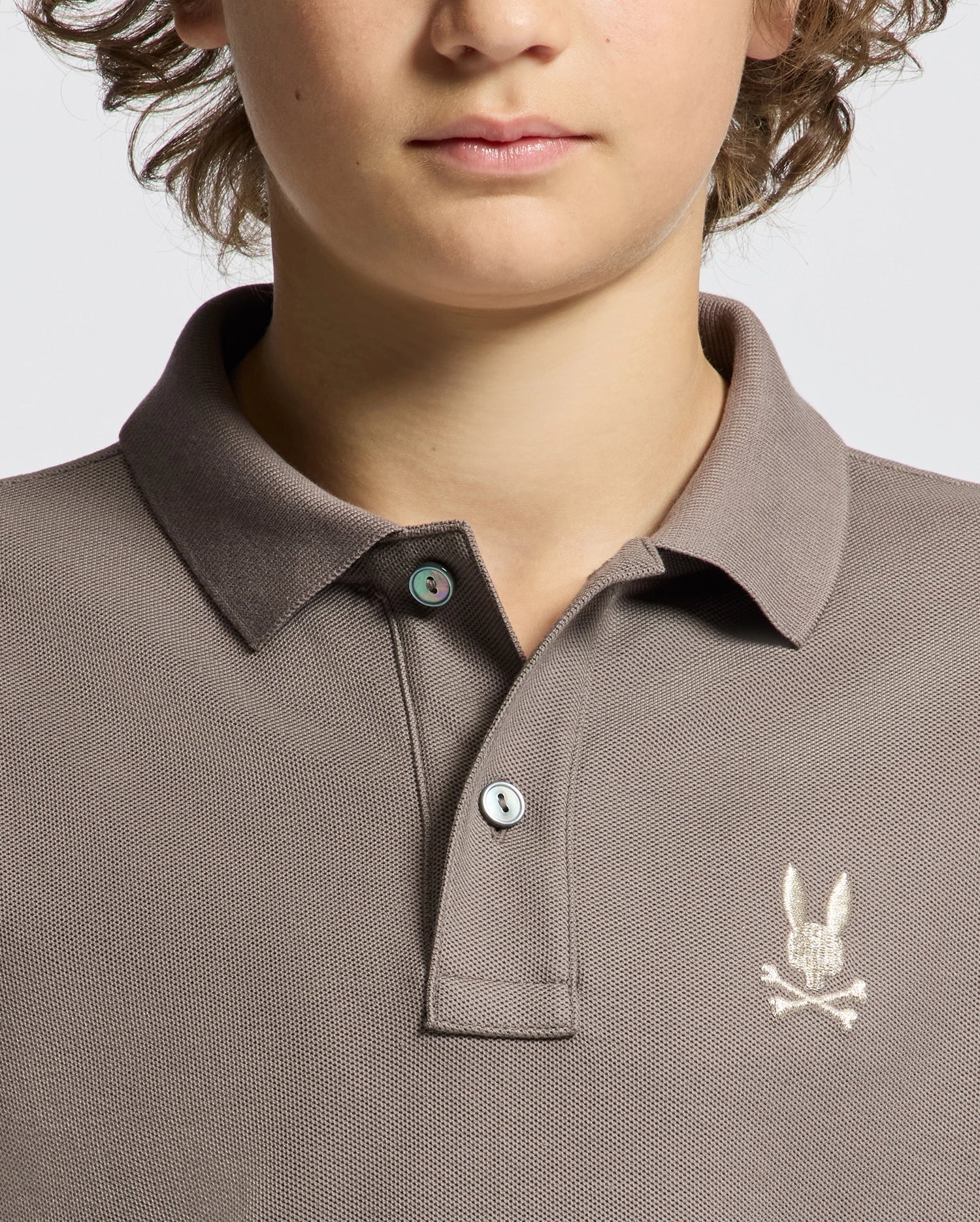 A person wearing a taupe KIDS ALEXANDER PIQUE POLO - B0K101E200 by Psycho Bunny, which features an embroidered rabbit skull and crossbones logo on the chest. This shirt is made from soft Pima cotton with two buttons and is depicted in close-up against a plain background.