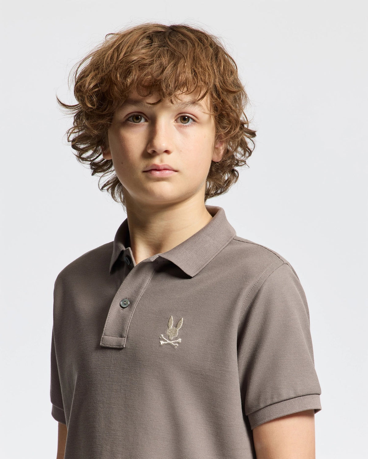 A young person with curly brown hair is wearing a gray Psycho Bunny KIDS ALEXANDER PIQUE POLO - B0K101E200, featuring an embroidered logo on the chest. This fashion-forward choice stands out against the plain white background.