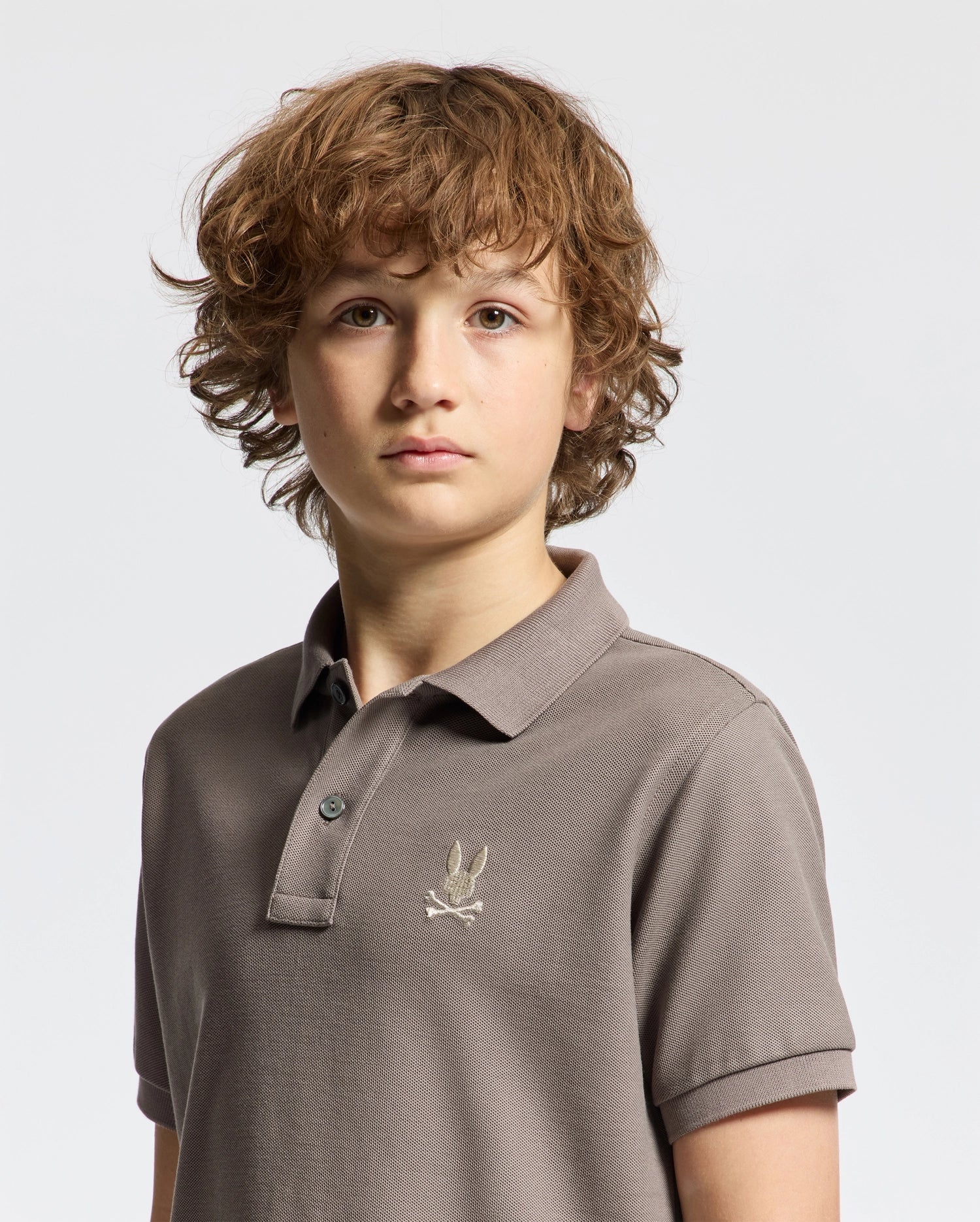 A young person with curly brown hair is wearing a gray Psycho Bunny KIDS ALEXANDER PIQUE POLO - B0K101E200, featuring an embroidered logo on the chest. This fashion-forward choice stands out against the plain white background.