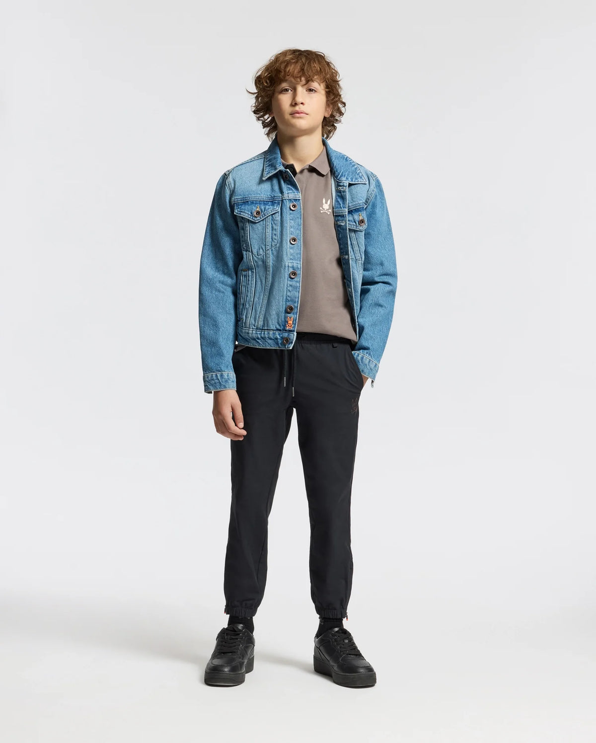 A young individual stands against a simple backdrop, showcasing a light blue denim jacket layered over a gray Psycho Bunny KIDS ALEXANDER PIQUE POLO (B0K101E200), accompanied by black pants and shoes. With their hands in their pockets and curly brown hair, they exude an effortless casual style.