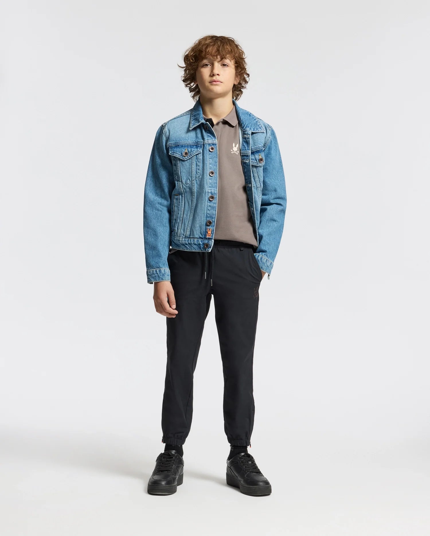 A young individual stands against a simple backdrop, showcasing a light blue denim jacket layered over a gray Psycho Bunny KIDS ALEXANDER PIQUE POLO (B0K101E200), accompanied by black pants and shoes. With their hands in their pockets and curly brown hair, they exude an effortless casual style.