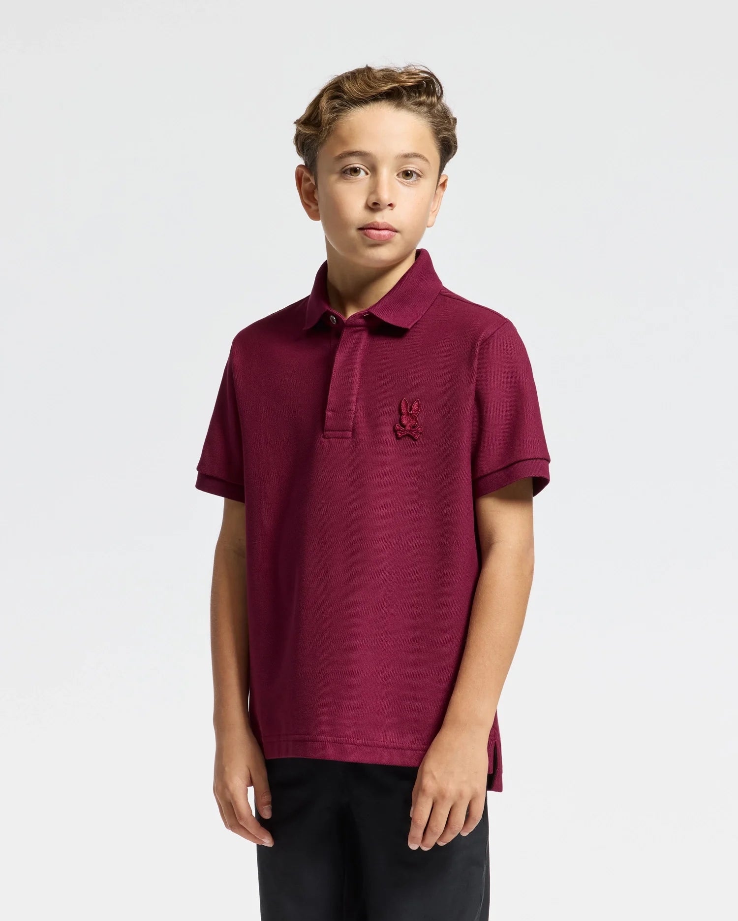 Against a light gray background, a young boy wearing the stylish KIDS BRANDSON PIQUE POLO - B0K108E200 by Psycho Bunny stands with short, wavy hair framing his expressionless face. The maroon polo, adorned with an embroidered logo, features a collar and short sleeves and is paired perfectly with his dark pants to create a classic look.