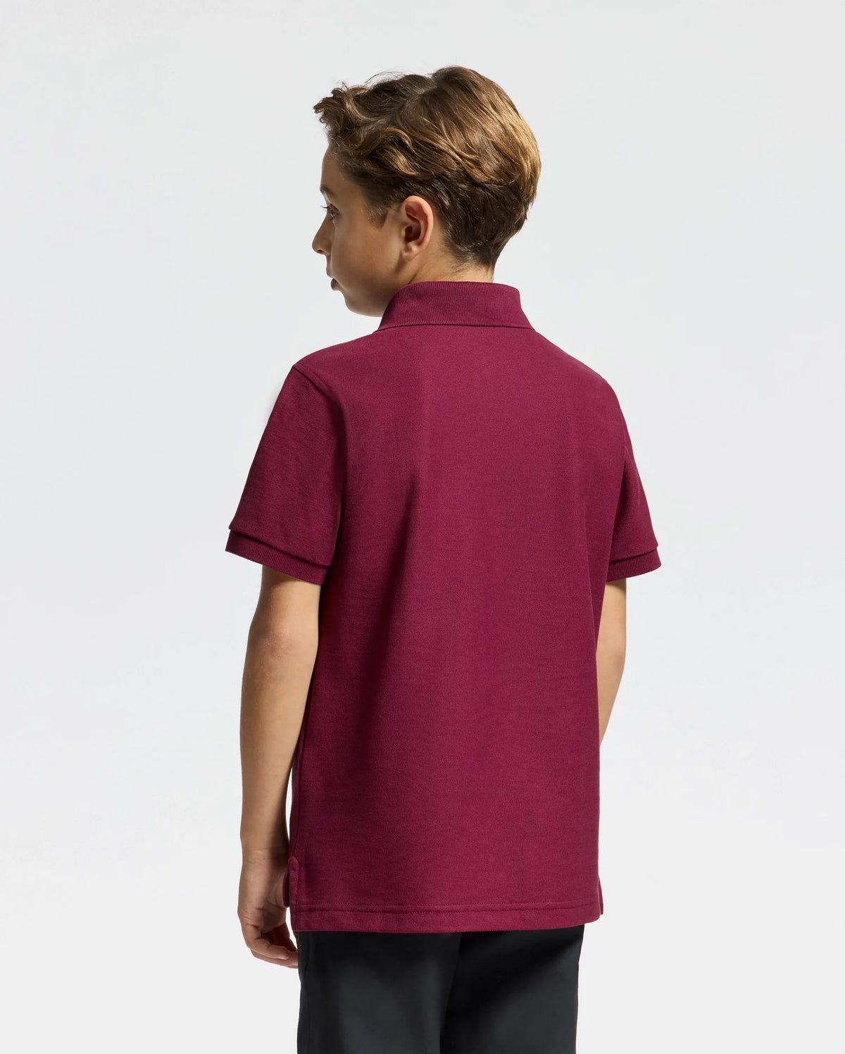 A young boy stands facing away, dressed in a stylish Psycho Bunny Kids Brandson Pique Polo in burgundy paired with dark pants. His hair is short and styled. The plain and neutral background accentuates the classic look of his soft Pima cotton polo.
