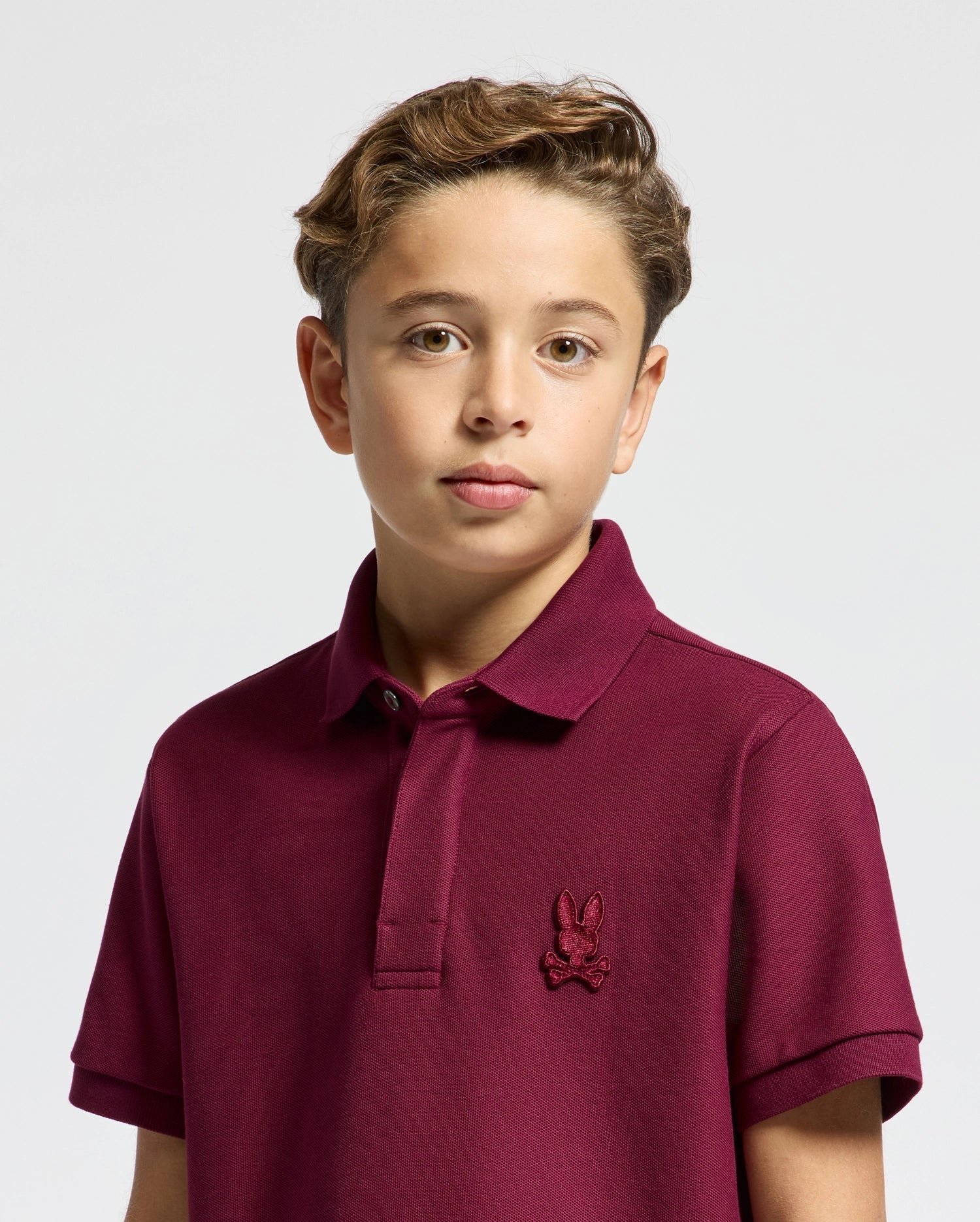 A young boy with short, light brown hair is seen wearing a stylish Kids' Brandson Pique Polo from Psycho Bunny in maroon, which features a small embroidered logo on the chest. He stands against a plain light gray background, giving his look an effortlessly elegant touch.