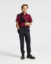 A young boy stands confidently against a plain background, dressed in the stylish Psycho Bunny KIDS BRANDSON PIQUE POLO - B0K108E200 in maroon, paired with black pants and shoes. A dark sports jacket is casually draped over his shoulder.