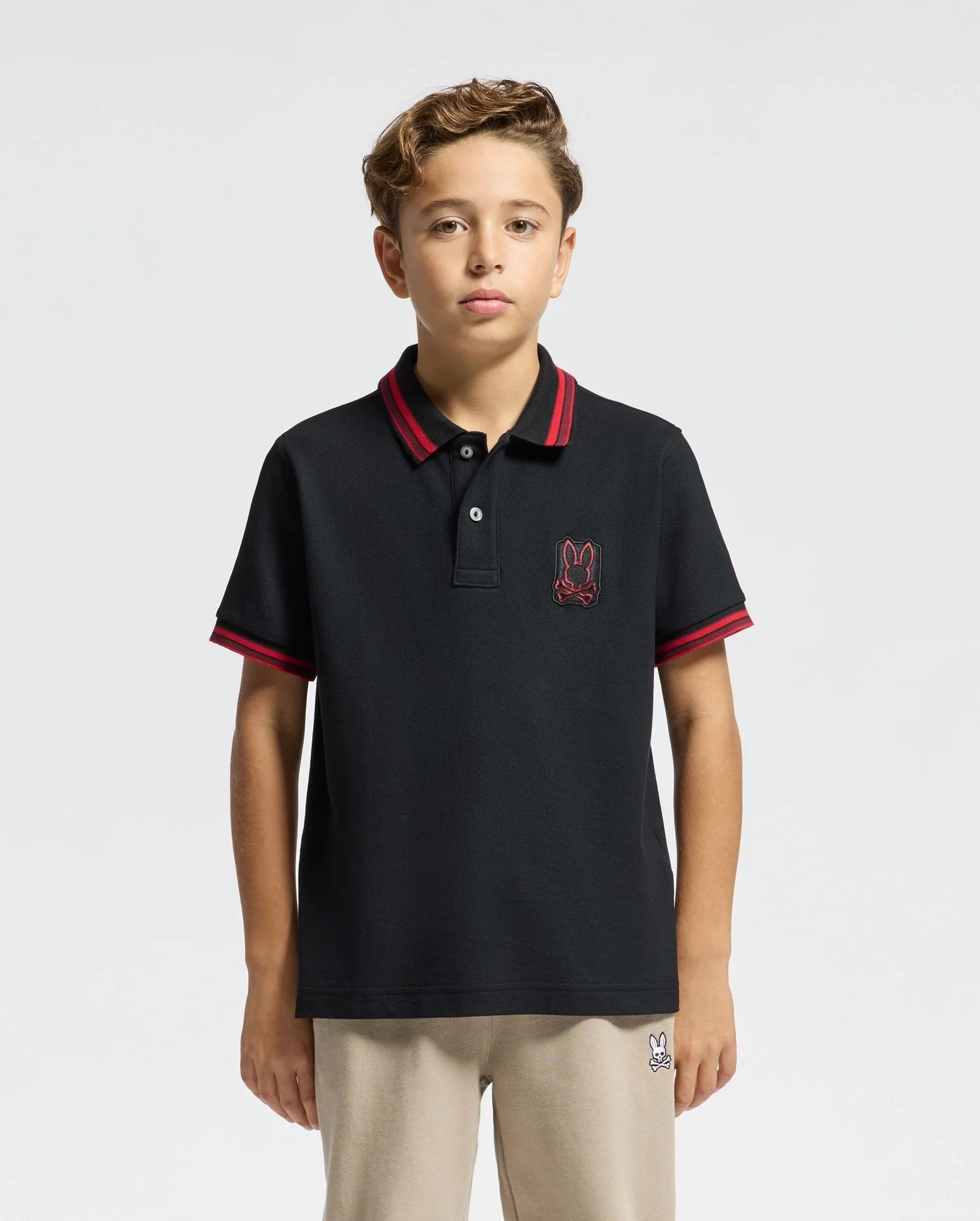 A young person with short curly hair is wearing the Psycho Bunny KIDS PIERCE PIQUE POLO - B0K201E200, which features premium quality material and red stripes on the collar and sleeves. This Pima cotton polo is adorned with an embroidered chest bunny logo. They are standing against a plain white background.